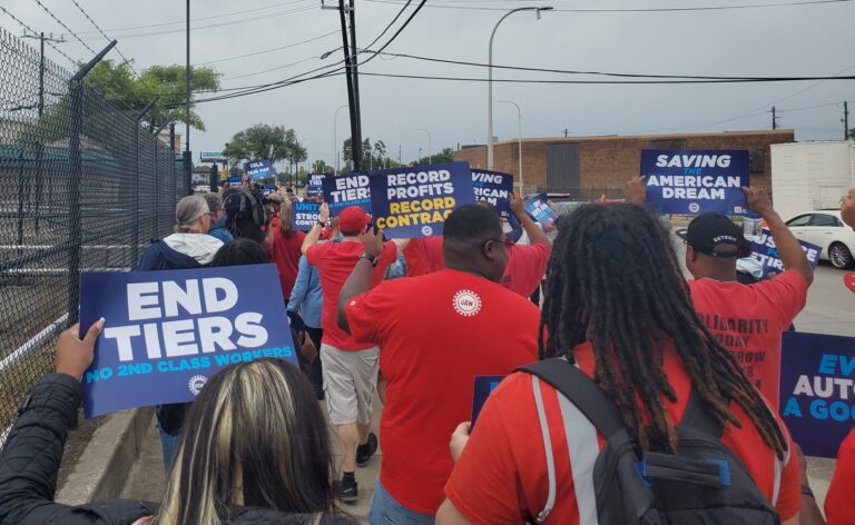 UAW members wearing red march on a picket line with their backs faced to the camera. Many of them are holding signs, which have slogans like "End Tiers: No Second Class Workers", "Record Profits, Record Contract", and "Saving the American Dream".