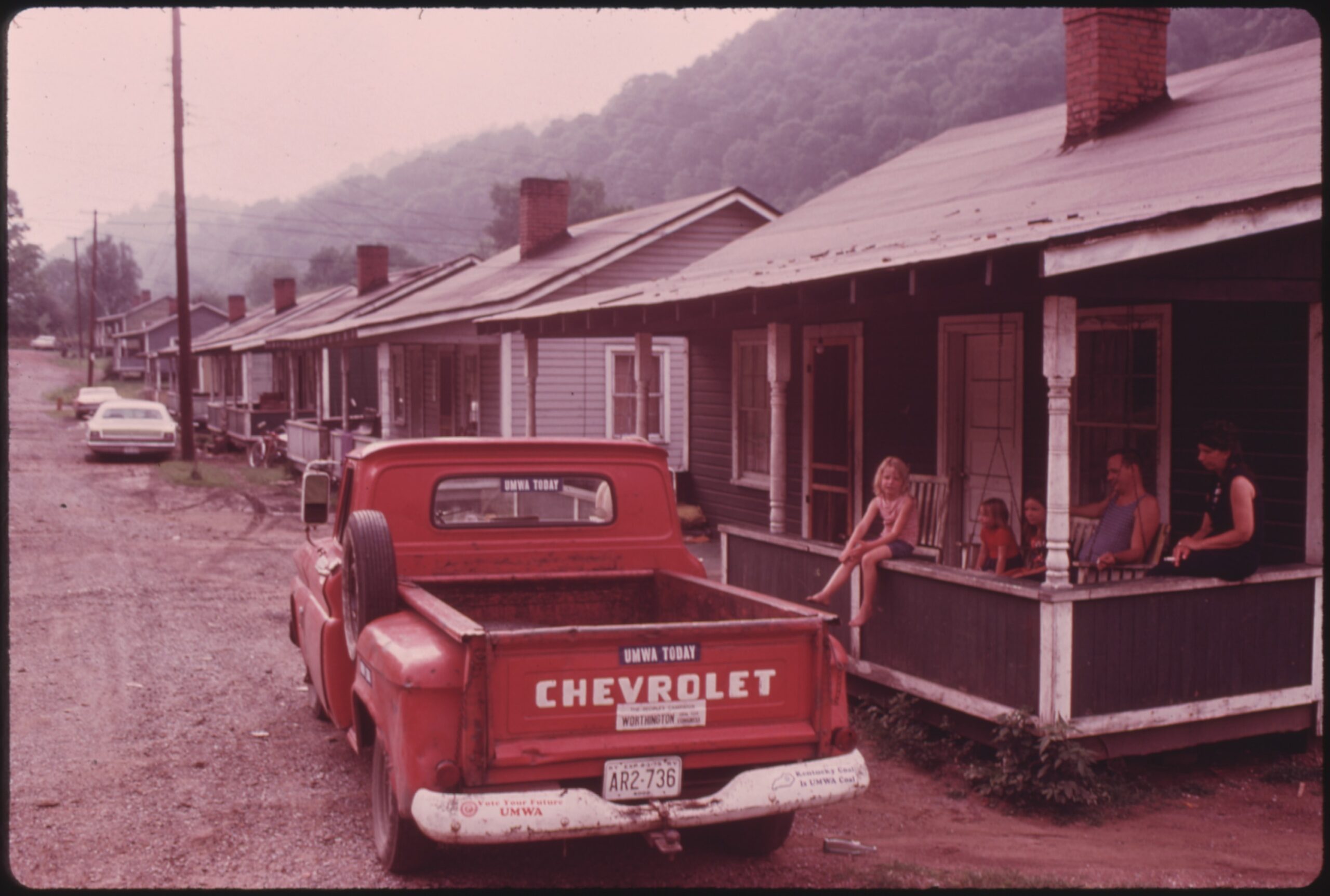 A red pick-up Chevrolet truck with stickers that say ‘UMWA TODAY,’ ‘Vote Your Future UMWA,’ and ‘Kentucky Coal is UMWA Coal,’ is parked in front of a row of small one-floor homes, all the same size, style and lay-out. The first home in view has three children and two adults sitting on the porch, looking out on the road and the car. The road that the houses sit on is gravel. Hilly forests are right behind the houses.