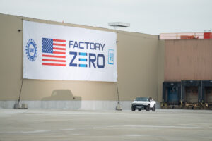 A white electric Hummer vehicle drives in an empty outdoor cement area of the General Motors plant Factory Zero. A large white flag covers one of the sides of the plant with text ‘FACTORY ZERO,’ the American flag, the UAW wheel logo, and the GM logo. Another part of the plant has dock doors for delivery trucks.