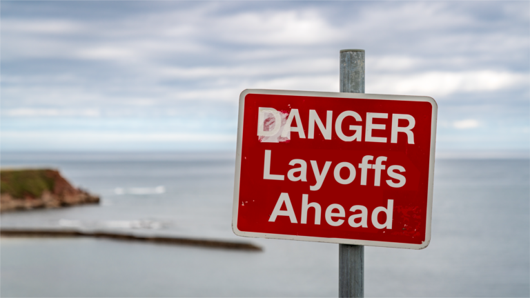 A red sign sits in the forefront of a photo with text ‘DANGER Layoffs Ahead.’ The background is cliffs overlooking a large body of water, either an ocean or a large lake.