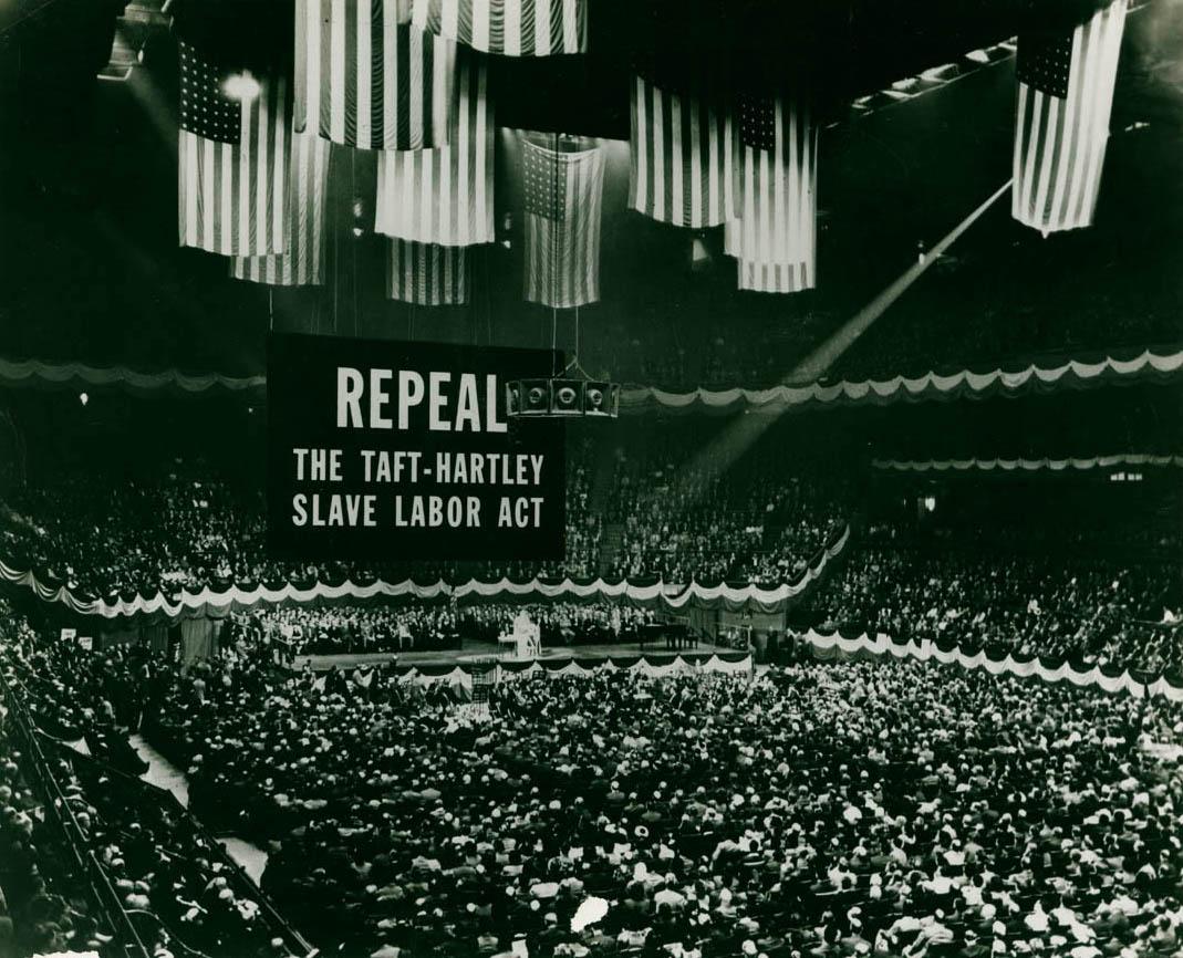 A black and white photo shows a large indoor arena filled with people. A large sign stands at one end of the arena with the text ‘REPEAL THE TAFT-HARTLEY SLAVE LABOR ACT.’ About eleven large American flags hang from the roof of the arena.