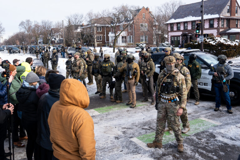 A photo shows about twenty ICE agents in the forefront, with more in the background, dressed in army garb and holding weapons, and with the majority having their faces masked, standing along a neighborhood street and facing off a group of community members wearing winter coats and hats. About 10 feet separates the two groups. The ICE agents in the forefront are standing on a bike lane. Snow covers the street and the yards and roofs of the houses.