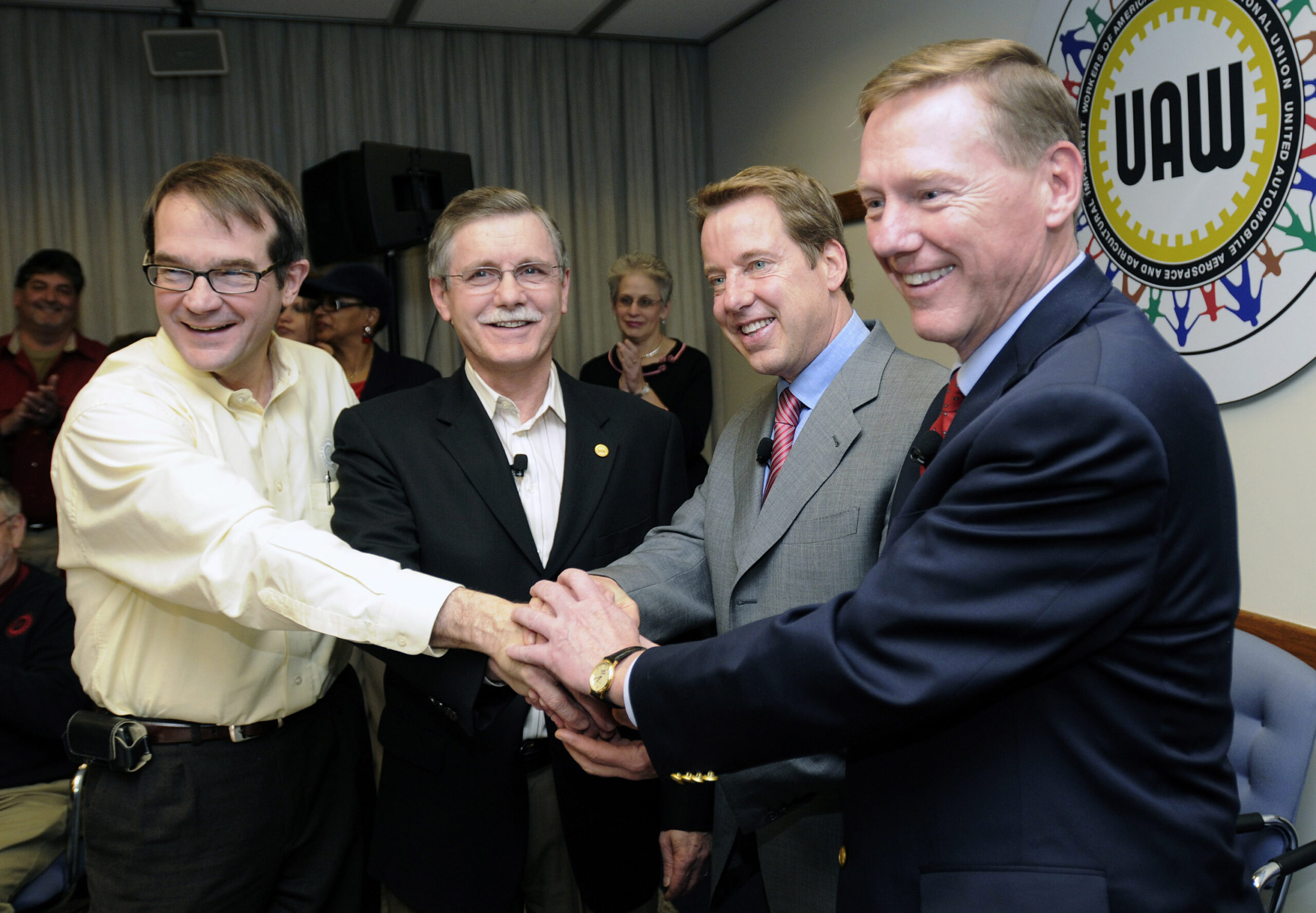 Four men in suits pose for a photo, smiling, with their hands embraced as a team. There is a small crowd of people in the background cheering them on. There is a large UAW wheel on a wall behind the men.