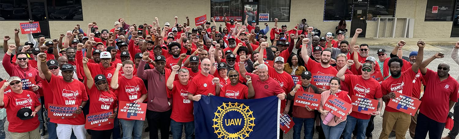 A large crowd of people smile and pose for a photo. They are all holding up power fists, and most are wearing red. Three people in the middle and the front hold up a blue flag with the yellow UAW wheel logo. People are also holding up signs that say ‘WE ARE ALABAMA WE ARE UAW’ and ‘MAKE MERCEDES A CAREER JOB.’