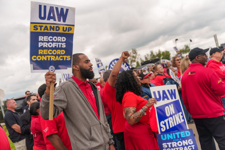 Man holding sign that says UAW Stand Up Record profits record contracts. Rally in the background with people holding fists