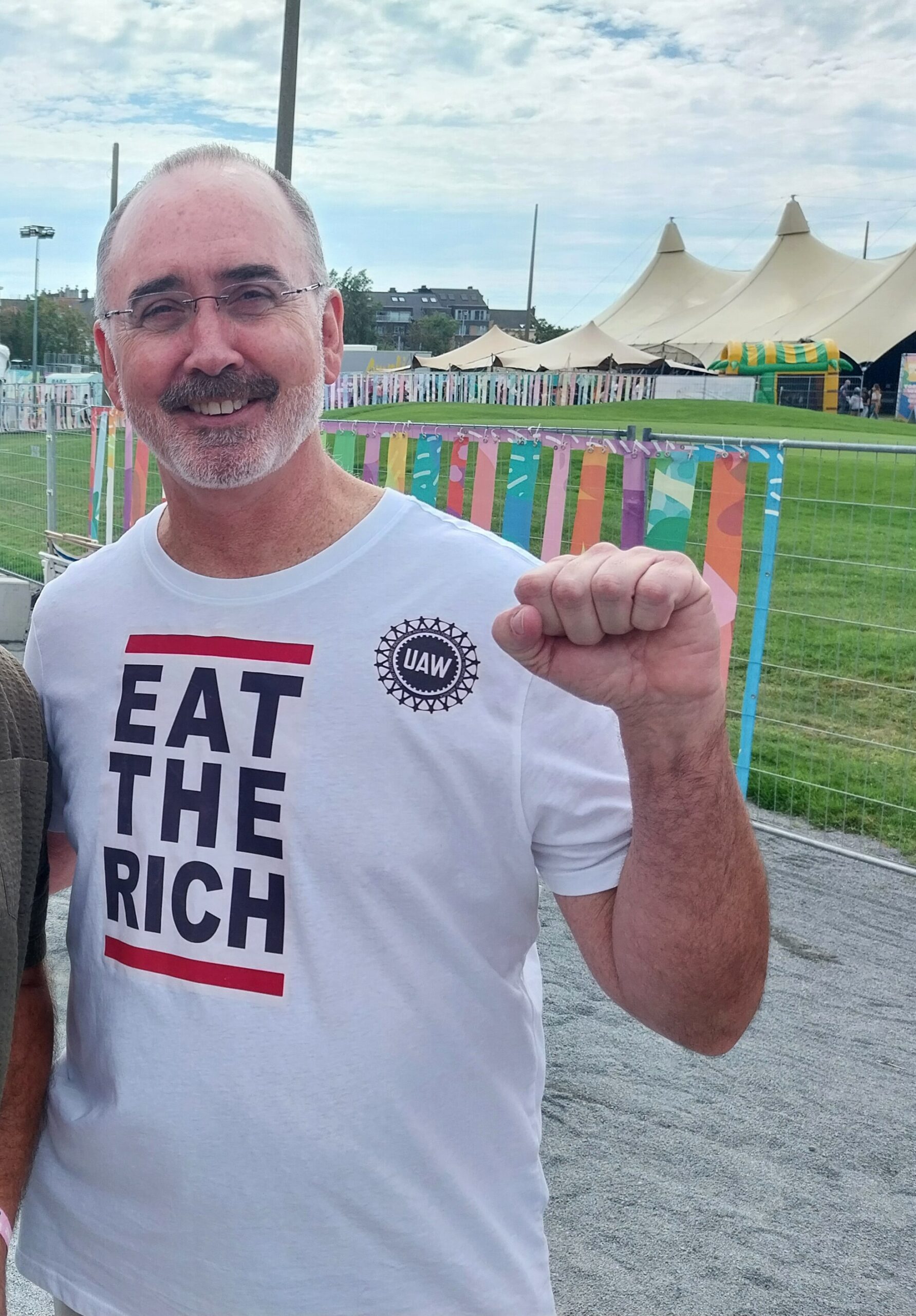 The older white man is Shawn Fain, he’s wearing glasses and stands in a white t-shirt printed with big bold text that says ‘EAT THE RICH.’ The t-shirt also has a United Auto Workers wheel logo near the left shoulder. Fain is smiling as he poses for the photo and holds up a power fist. There are decorations in the background for an outdoor festival.
