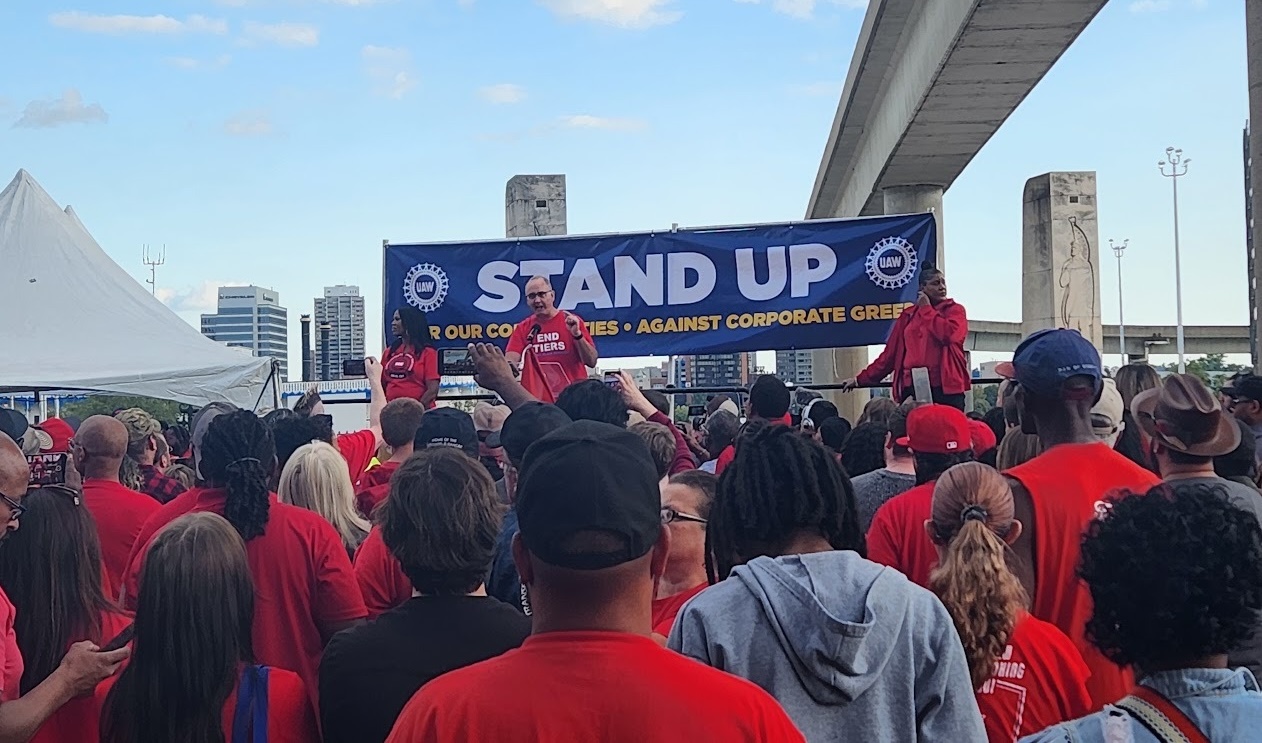 The photo catches the backs of a crowd outside facing a stage with speakers. The middle speaker is a man speaking into the microphone, with a shirt that says ‘END TIERS.’ The left and right speakers are women looking off to the side. Behind them is a large sign that says ‘STAND UP AGAINST CORPORATE GREED,’ with two of the Unite All Workers for America wheel logo. There are some clouds in the sky.