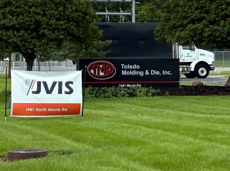 This is an image of a truck behind the sign for "Toledo Molding & Die, Inc." There is grass in the foreground and trees in the background.