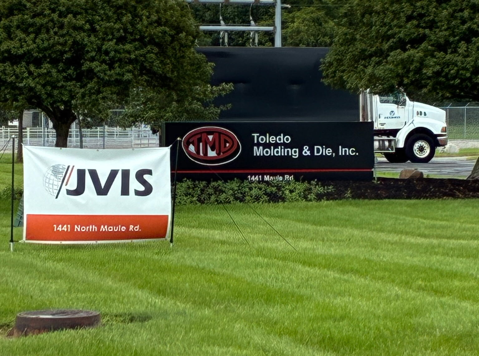 This is an image of a truck behind the sign for "Toledo Molding & Die, Inc." There is grass in the foreground and trees in the background.