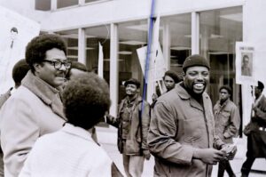 A group of smiling Black workers are protesting outside. They are wearing warm clothes and holding picket signs.