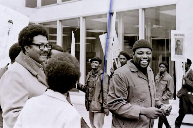 A group of smiling Black workers are protesting outside. They are wearing warm clothes and holding picket signs.