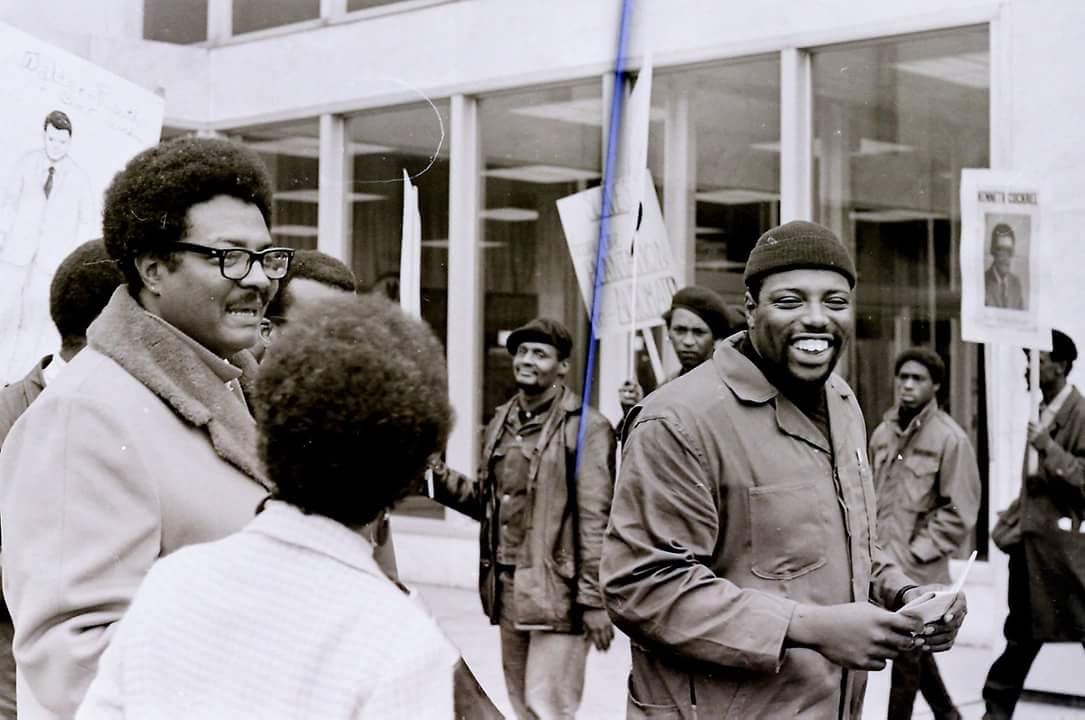 A group of smiling Black workers are protesting outside. They are wearing warm clothes and holding picket signs.
